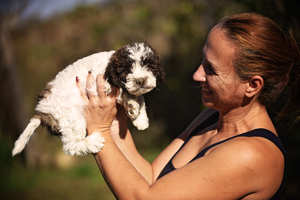 Biljana Bjekić drži štene u rukama, oboje izražavaju radost i zadovoljstvo, simbolizujući bliskost i ljubav između odgajivača i šteneta, i predstavljaju odgovoran odgoj Lagotto Romagnolo